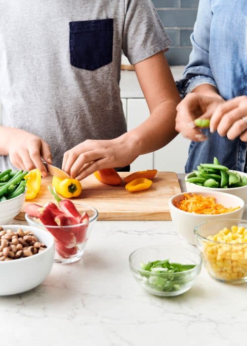 Two kids cutting fresh vegetables on a wooden cutting board with ingredients in bowls in a beautiful white marble kitchen