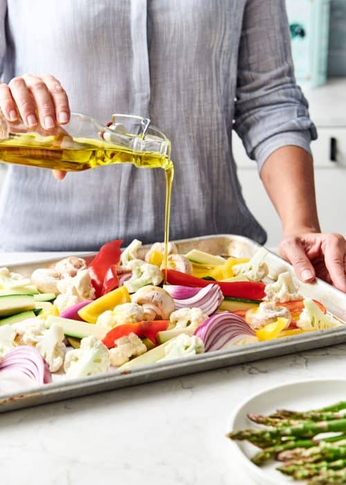 Woman pouring olive oil on a baking sheet filled with an assortment of vegetables on a white marble countertop