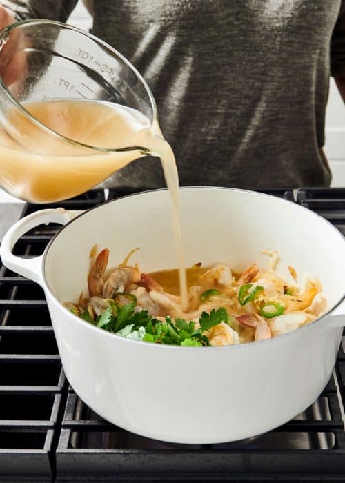 Pouring a rich resturant style broth into a Le Creuset Dutch oven filled with shrimp and other ingredients on a metal cooktop