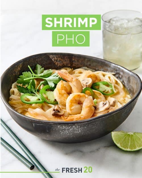 Black ceramic soup bowl filled with suculent shrimp pho with lime wedge, chopsticks & a glass of water on a white marble surface