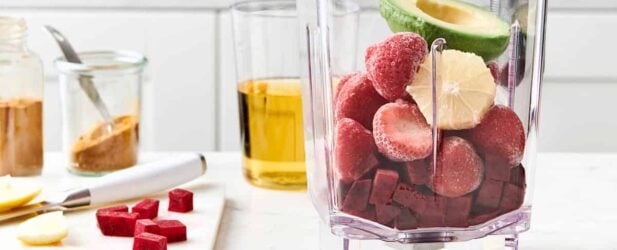 Blender full of beets, strawberries & avocado with ingredients on a wood cutting board in a white marble kitchen