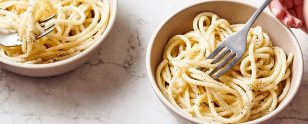 2 kids eating Italian cacio e pepe pasta from 2 bowls with chili flakes & lemon water on a white marble surface