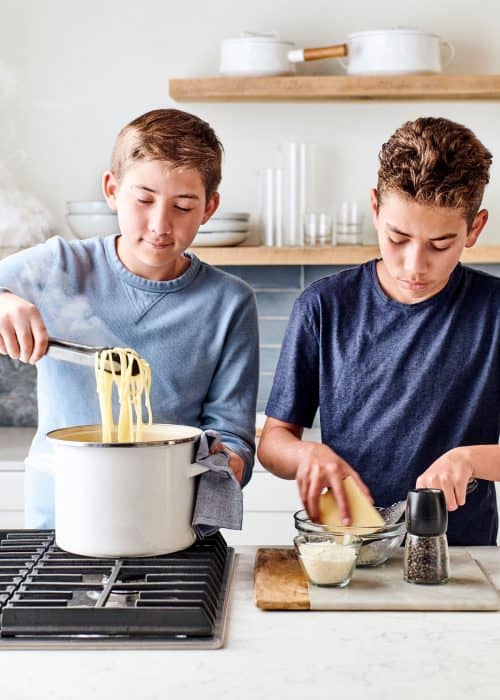 Two boys cooking pasta noodles in a white stockpot & grating parmesan cheese in a white marble kitchen