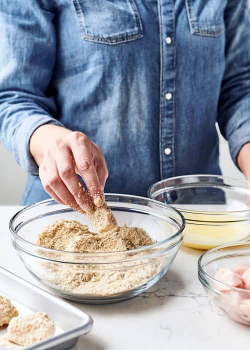 Woman breading chicken nuggets in a glass bowl of bread crumbs, parmesean & egg wash on a white marble surface