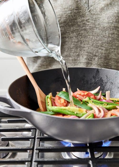 Pouring water into a Le Creuset wok filled with chilis, onions & tomatoes on a gas cooktop