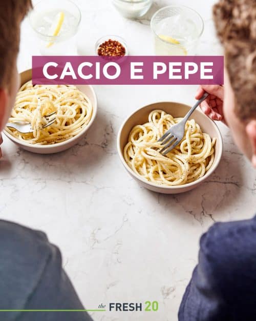 2 kids eating Italian cacio e pepe pasta from 2 bowls with chili flakes & lemon water on a white marble surface