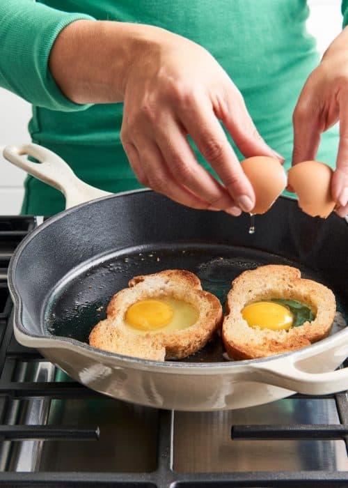 Woman cracking two eggs into two peices of hollowed out toast on a Le Cruset pan on a metal stovetop