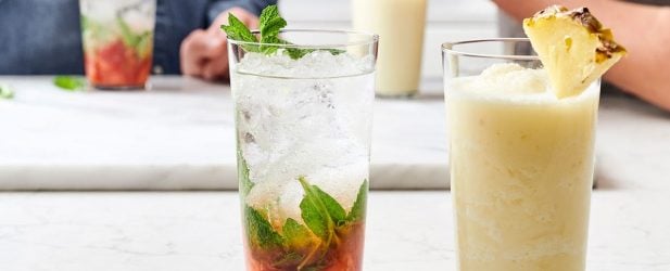 2 boys making 2 fresh fruit summer mocktails with mint leaves & pineapple garnish in a white marble kitchen