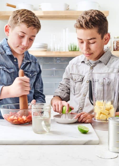 2 young boys muddling strawberries & juicing limes with a pitcher of pineapple in a white marble kitchen