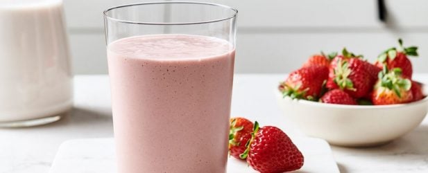 Glass of no sugar added strawberries & cream smoothie with a bowl of strawberries on a white marble surface in a beautiful kitchen