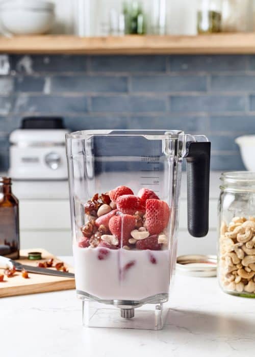 Blender full of strawberries, almond milk, nuts, dates & other ingredients on a wood cutting board in a white marble kitchen