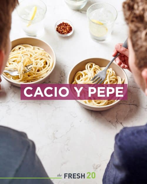 2 kids eating Italian cacio e pepe pasta from 2 bowls with chili flakes & lemon water on a white marble surface