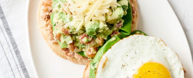 Oval plate with 2 summer style guacamole tostadas with lettuce, cheese and sunny side up egg on a white marble surface with a linen napkin