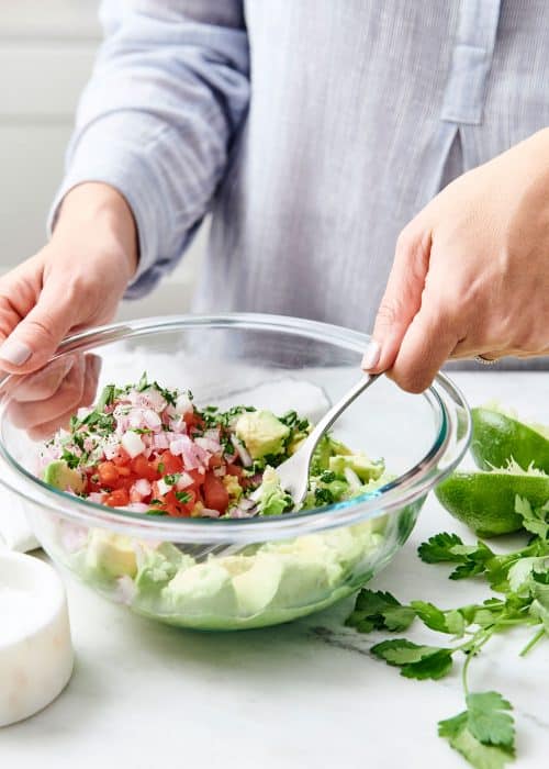 Woman mashing avocados, tomatoes & onions with a fork in a glass bowl for guacamole with lime and cilantro on a white marble surface