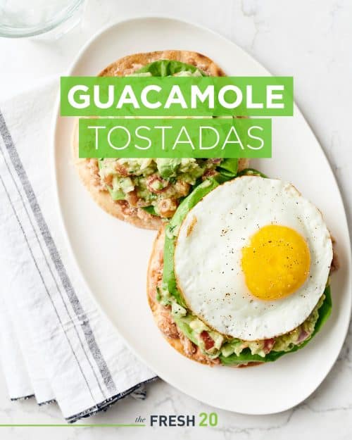 Oval plate with 2 summer style guacamole tostadas with lettuce, cheese and sunny side up egg on a white marble surface with a linen napkin