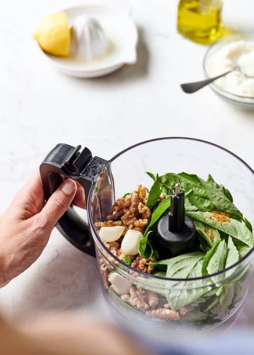 Food processor full of fresh basil, garlic and walnuts along with a lemon juicer & olive oil on a white marble surface