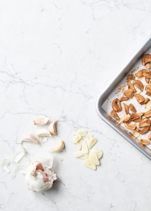 Baking sheet full of crispy baked garlic alongside raw chopped garlic cloves on a white marble surface