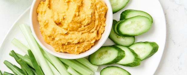 Ceramic plate filled with cucumber slices, green beans & celery with a bowl of spicy hummus on a white marble surface