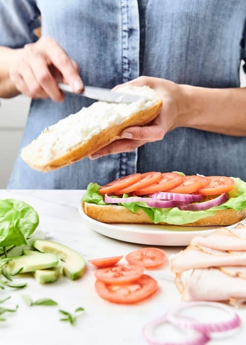 Woman spreading mayo on a layered turkey sandwich next to a variety of fresh ingredients on a white marble surface
