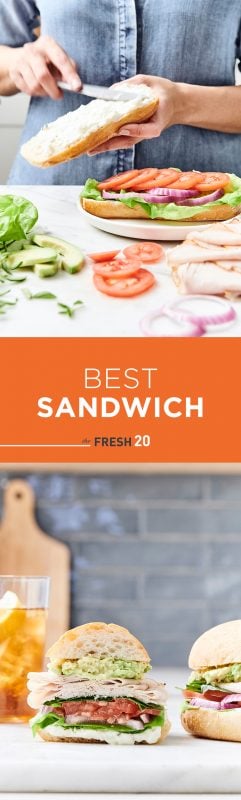 Woman spreading mayo on a layered turkey sandwich next to a variety of fresh ingredients in a white marble kitchen