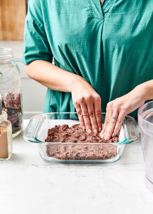 Woman pressing her hands into no bake breakfast bar batter into a glass baking dish next to jars of ingredients in a beautiful white marble kitchen