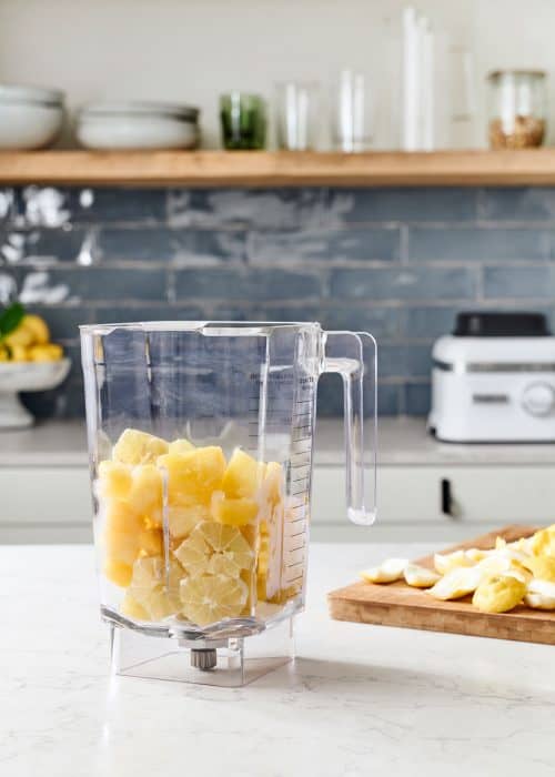 Blender full of yellow lemons & pineapple in a beautiful marble kitchen with a wood cutting board with fruit