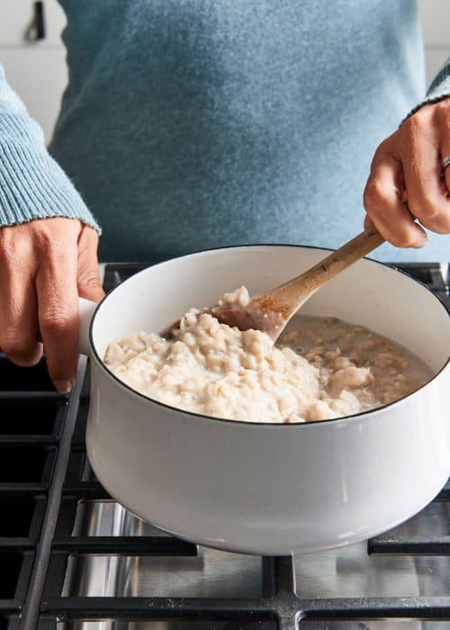 Woman stirring a fresh batch of oatmeal in a white pot on a black metal cooktop