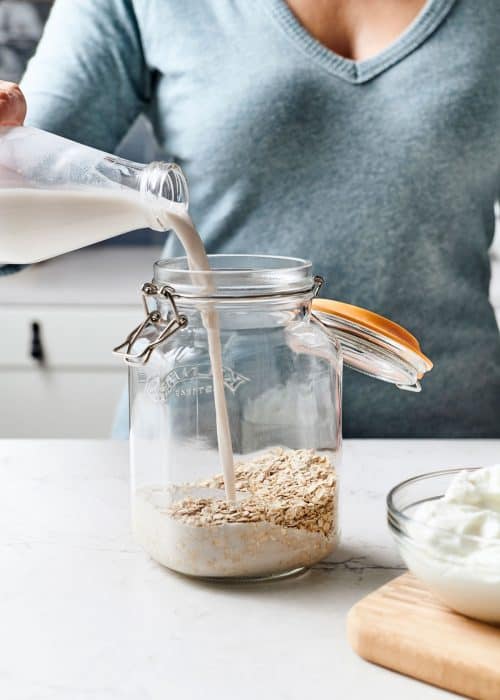Woman pouring fresh almond milk into glass mason jar filled with oatmeal in a white marble kitchen