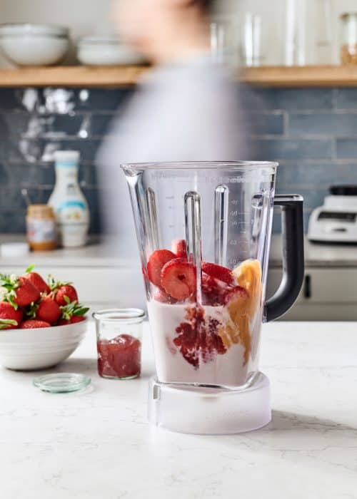 Blender full of almond milk, peanut butter, & a bowl of strawberries & jam in a white marble kitchen