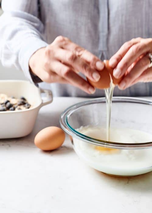 Woman cracking eggs into a glass mixing bowl filled with milk cinnamon & vanilla on a white marble surface