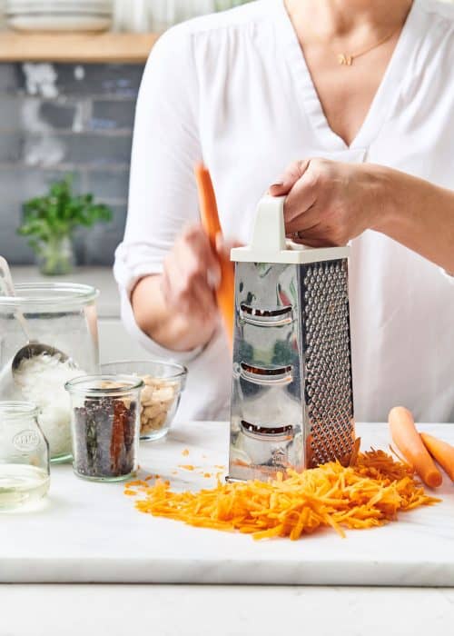 Woman shredding carrots with a metal box grater onto a white marble surface with raisins, coconut & nuts