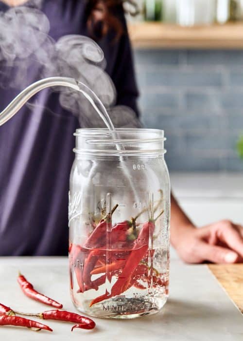 Woman pouring steaming boiling water from a kettle into a mason jar filled with deep red hot chili peppers