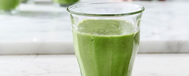 Woman making a glass of morning kale grape mint smoothie in a white marble kitchen