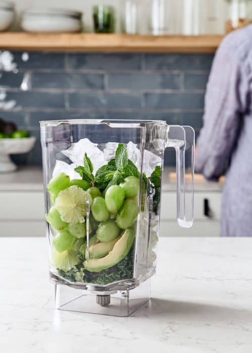 Woman filling a blender full of green grapes, mint, kale, lime & avocados in a white marble kitchen