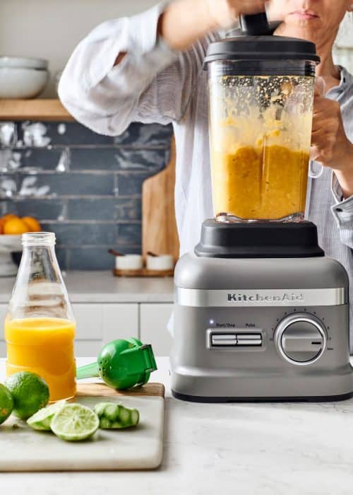 Woman using KitchenAid blender filled with mangos, limes and orange juice in a white marble kitchen