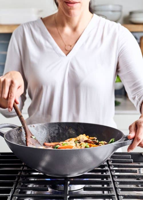 Woman stirring a Le Creuset gray wok filled with 8 vegetable stir-fry on a cooktop in a white marble kitchen