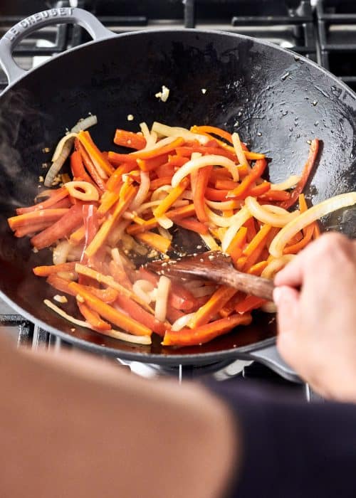 Le Creuset wok filled with carrots, onion and bell peppers being stirred with a wooden spatula on a cooktop