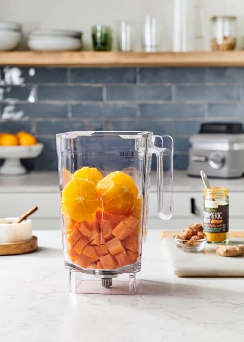 Blender full of oranges & carrots with ginger & turmeric on a white marble counter in a modern kitchen