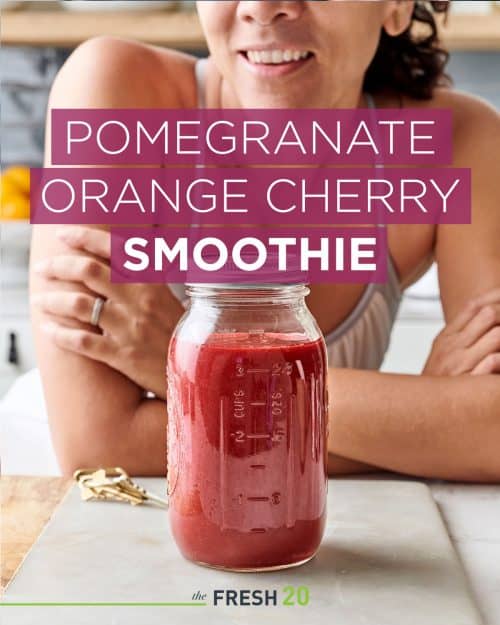 Woman leaning behind a mason jar filled with festive holiday pomegranate orange cherry smoothie on a white marble surface