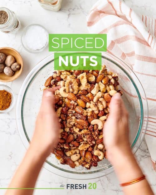 Hands mixing spiced nuts in a glass bowl on a white marble surface for the holidays