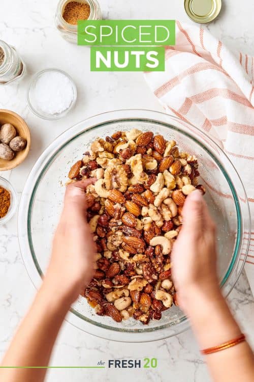 Hands mixing spiced nuts in a glass bowl on a white marble surface for the holidays