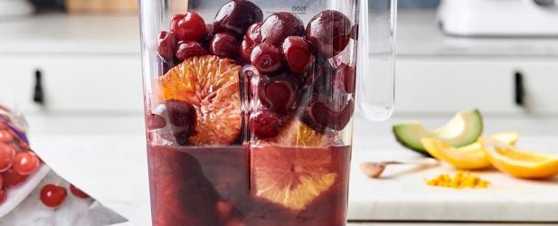 Blender full of pomegranate juice, oranges, cherries & avocado on a white marble surface in a beautiful kitchen