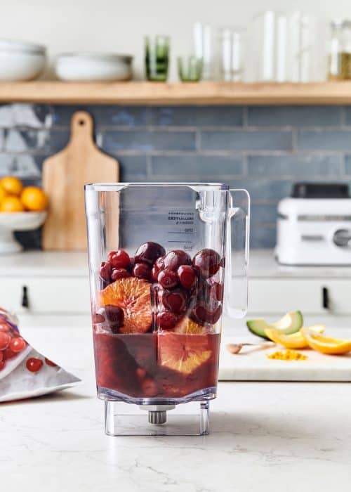 Blender full of pomegranate juice, oranges, cherries & avocado on a white marble surface in a beautiful kitchen