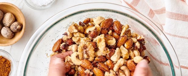 Hands mixing spiced nuts in a glass bowl on a white marble surface for the holidays