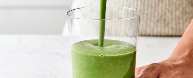 Woman pouring green banana chai spice superfood smoothie into a clear glass in a white marble kitchen