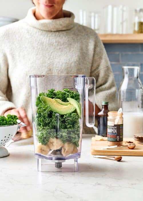 Woman making a blender full of kale, avocados, bananas & chai spices in a white marble kitchen