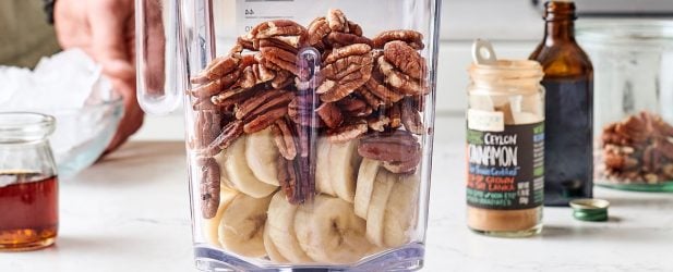 Man making a blender full of bananas, pecans, cinnamon, honey & vanilla in a white marble kitchen