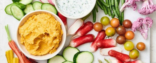 A white marble slab filled with an assortment of dipping vegetables along with three bowl of flavorful crudites