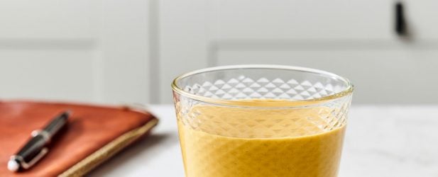 Glass filled with creamy carrot cake smoothie with a leather notebook & pen in a white marble kitchen