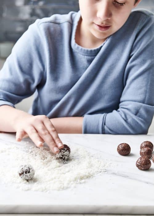 Young boy rolling heart healthy raw chocolate truffles in a pile of shredded coconut in a white marble kitchen